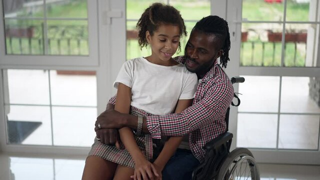 Portrait of beautiful teenage African American girl sitting on lap of handsome disabled man in wheelchair talking smiling. Front view positive happy daughter and father enjoying weekend leisure