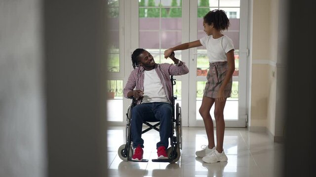 Wide shot cheerful teenage girl dancing in slow motion with disabled man in wheelchair. Portrait of happy carefree African American daughter having fun with father at home in living room