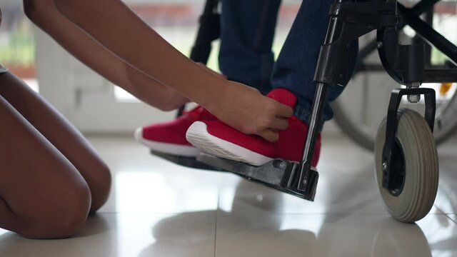 Teenage African American female hands tying shoelaces on red sneakers of disabled man in wheelchair in slow motion. Unrecognizable empathetic daughter taking care of father at home indoors