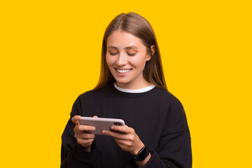 Cheerful girl gamer playing on mobile phone and smiling over yellow background