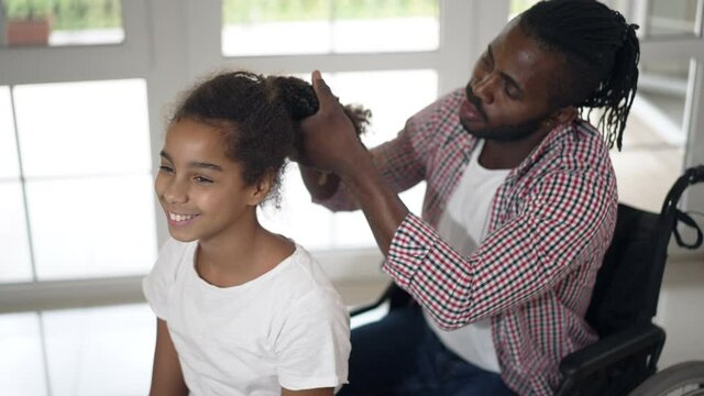 Charming happy teenage African American girl smiling talking as disabled man in wheelchair making ponytail. Portrait of daughter chatting with father helping with hairdo at home indoors