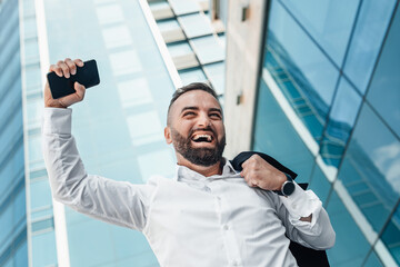 Mature job seeker celebrating successful interview raising fist with smartphone walking against glass business center