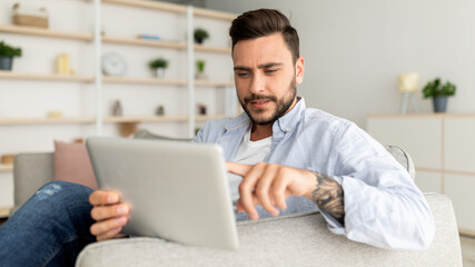 Closeup of handsome man reclining on sofa, using modern pad at home interior, resting on couch in living room