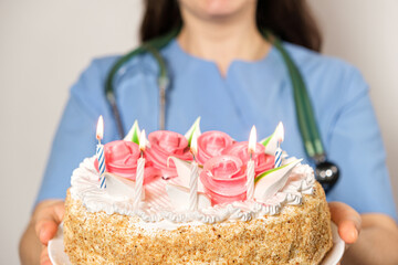A woman doctor in a blue uniform holds a birthday cake with candles and smiles.