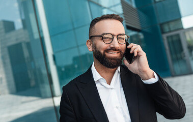 Cheerful middle-aged employee talking on smartphone and smiling while walking near office center, free space