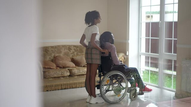 Side view teenage daughter talking in slow motion with disabled father in wheelchair looking out the window at home. Wide shot shooting through glass positive girl and man chatting in living room