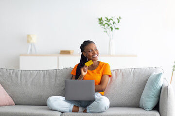 Young African American woman buying online with credit card and laptop, sitting on couch at home,...