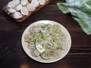 Boiled dumplings with butter, spices and green onions in a plate, frozen dumplings on a kitchen board, napkin on a dark wooden table, flat layout, top view. Traditional meat dish with dough