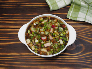 Traditional okroshka soup with vegetables and kvass in a white bowl on a wooden table, flat layout Recipe for cold spring soup with ridis, cucumber for diet food