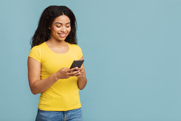 Cool trendy gadget. Excited african american lady using smartphone, standing over blue background with copy space