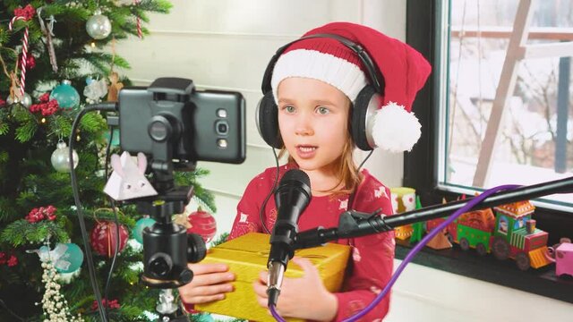 a girl in headphones and Red Santa Claus Hat is singing into a microphone near a Christmas tree and holding a gift in her hands, looking at the smartphone screen. High quality 4k footage