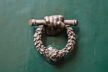 Close-up of an old green door with a bronze knocker representing a hand holding a laurel wreath...