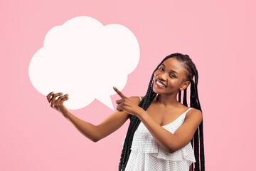Cheerful black woman with afro bunches pointing at blank speech bubble on pink studio background,...