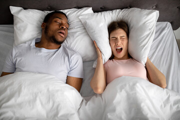 Top view of snoring black husband and his stressed wife screaming and covering ears with pillow in...
