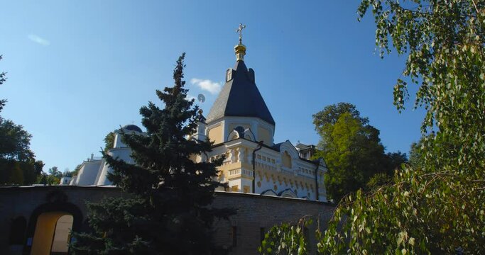 A beautiful view of the Orthodox Christian Church in the monastery, built on a hill covered with conifers on a sunny day, against the backdrop of a cloudless sky.