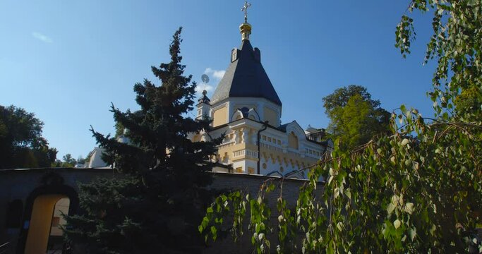 A beautiful view of the Orthodox Christian Church in the monastery, built on a hill covered with conifers on a sunny day, against the backdrop of a cloudless sky.