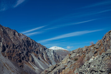 Colorful alpine landscape with great mountain in autumn colors with snow on peak under cirrus clouds in blue sky. Awesome autumn scenery with rocks and snow-covered mountain top. Scenic mountain view.