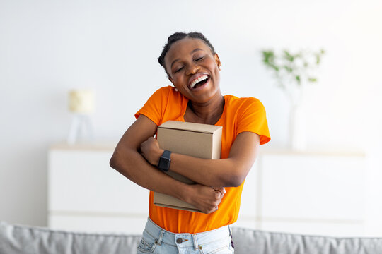 Happy black woman hugging cardboard parcel, receiving desired product, getting online order delivered at home