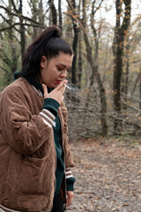 Fototapeta na wymiar An addict young woman is smoking in forest. Harmful habit. Dangerous cigarette. The concept of 