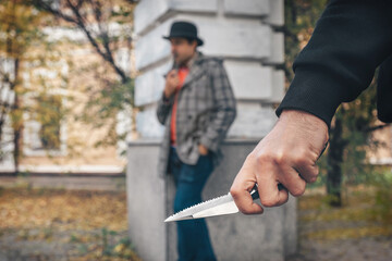 Hand of a robber with a knife on the background of a man in the park. Crime, violence concept