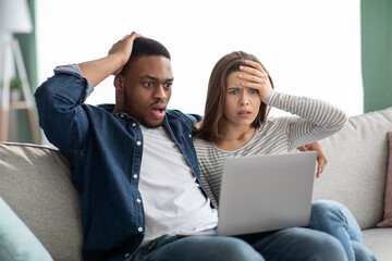 Shocking News. Terrified multicultural couple looking at laptop screen and touching heads