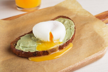 Healthy avocado toast for breakfast with rye bread, poached egg served on wooden board. Orange juice on the background. Clean eating.
