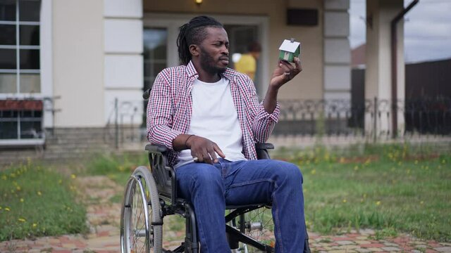 Portrait of African American disabled man in wheelchair outdoors looking at toy house with blurred teenage girl jumping in slow motion at background on porch. Thoughtful ill guy planning home selling
