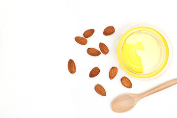 Yellow almond oil in a clear bowl with a wooden spoon next to it and several almonds on a white background