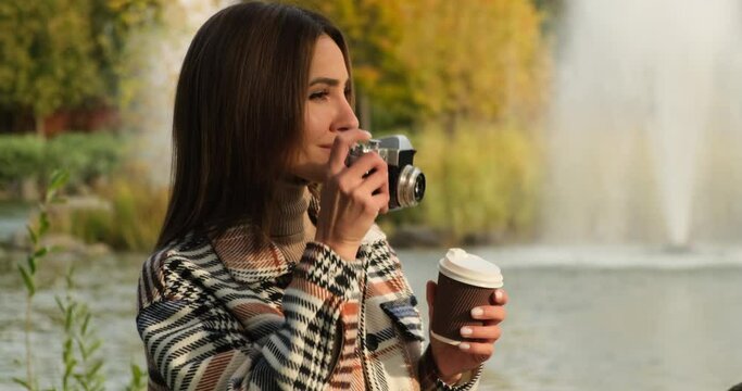Beautiful woman drink coffee and posing for the camera in autumn park. a female in a plaid coat is drinking tea or coffee in a warm autumn park, waiting for her boyfriend on a date. fountain, birds