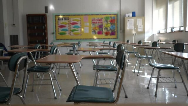 Desks in empty dark high, middle, or elementary school classroom with light coming through windows.
