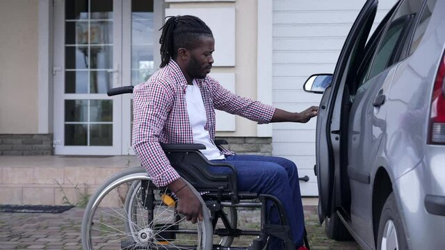 Side view African American man rolling wheelchair to car opening driver's seat door. Portrait of motivated confident disabled driver outdoors on front yard at vehicle. Lifestyle and accident concept