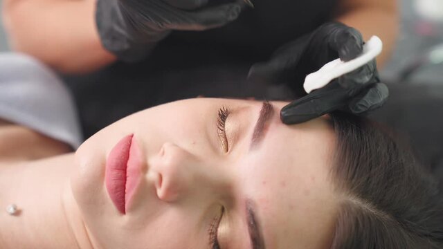 Master makes permanent eyebrow makeup procedure using special needle tattoo machine to woman in beauty salon. Microblading brows tattooing. Dark pigment is injected under skin. Cosmetology procedure