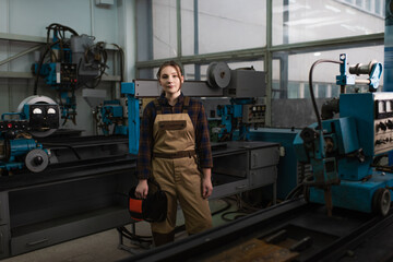Fototapeta premium Brunette welder holding protective helmet near welding machines in factory