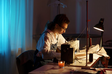 Busy tailor woman working late at night sit at table with sewing machine, fabric and paper patterns in studio creating new collection for small atelier opening. Tailoring entrepreneurship concept