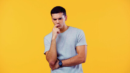 pensive young man in t-shirt touching lips isolated on yellow.