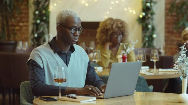 Young African American man sitting at cafe table with glass of wine on it and typing on laptop