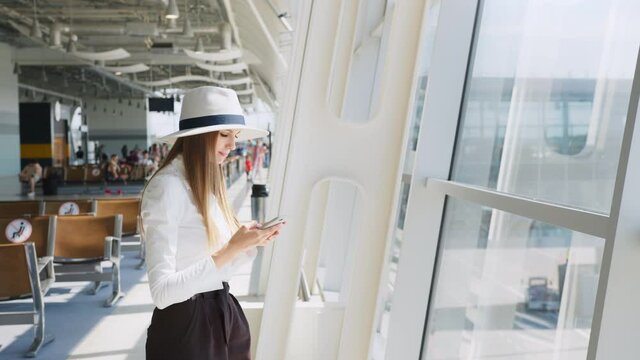 Girl uses smartphone in the airport lounge. Beautiful young girl using smartphone communication application airport arriving in new country. Woman travels around world. Smiling at the camera.