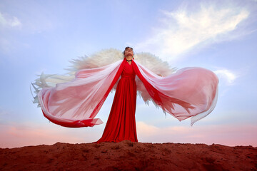 Beautiful young woman or girl with curly hair and in red dress with a light flying fabric and white wings on the sand on sunny day with blue sky. Angel model or dancer posing in photo shoot on dunes
