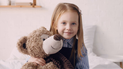 Positive kid hugging teddy bear and looking at camera in bedroom.