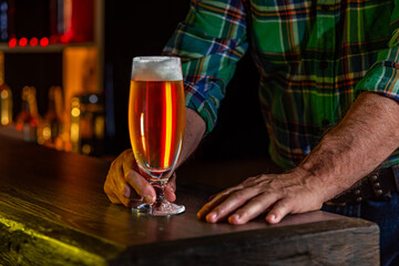 Hands of a senior male barman, bartender, pantryman. Glass of beer, ale on the wooden vintage bar counter. Holiday of Ireland on St. Patrick's Day in irish pub, bar