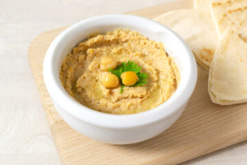 Hummus in a ceramic white bowl with pieces of pita on a wooden board on a light background. A traditional Middle Eastern dish. Horizontal orientation. Vegetarian food concept.
