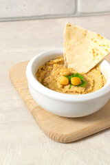 Hummus in a ceramic white bowl with pieces of pita on a wooden board on a light background. A traditional Middle Eastern dish. Horizontal orientation. Vegetarian food concept. Vertical orientation.