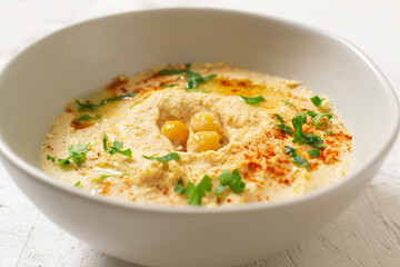 Hummus in a ceramic white bowl with paprika and parsley leaves on a light background. A traditional Middle Eastern dish. Horizontal orientation. Vegetarian food concept. Close up