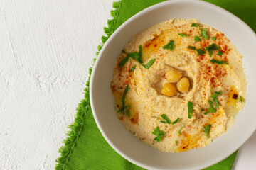 Hummus in a ceramic white bowl with paprika and parsley leaves on a green napkin. A traditional Middle Eastern dish. Horizontal orientation. Vegetarian food concept. Top view
