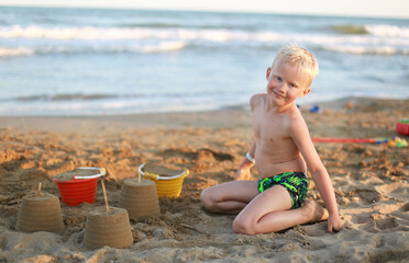 A blonde smiling 5 years old child boy on the beach building sand castles