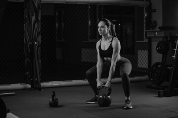 Fototapeta na wymiar A sporty woman with dark hair is squatting with a kettlebell in a gym. A girl in shorts is doing a workout with an iron kettlebell.