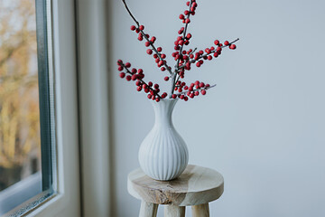 Scandinavian interior design of living room with stylish vase with red ilex against wall and large window. Copy space. Minimal concept. DIY apartment decor. Winter decoration.