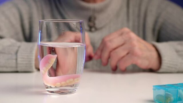 Elderly woman takes false teeth out of her mouth and puts it in glass of water. Oral, denture and dentures hygiene.