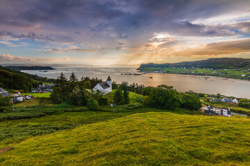 Fototapeta na wymiar a place in Scotland on the Isle of Skye. Church on a hill with green meadow and trees. View over the landscape with sun rays on the horizon. Dramatic clouds in the sky at sunset