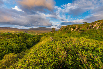 Landscape on the Isle of Skye of Scotland. Sunshine in summer with green meadows, hills, bushes and shrubs. Single rock spire grom Castle Ewen in the background. Blue sky with clouds